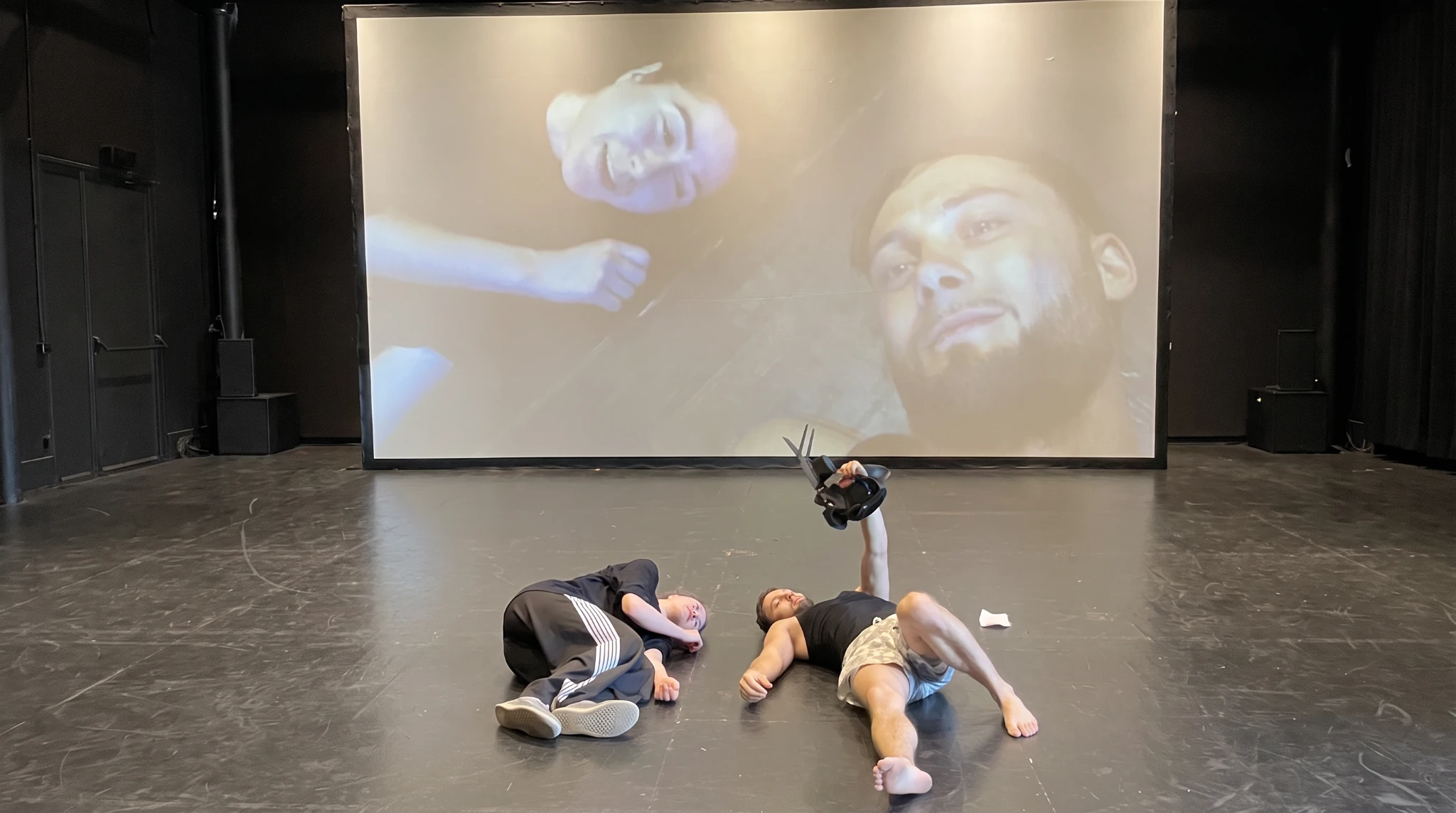 Deux interpr&egrave;tes allong&eacute;s au plateau, une cam&eacute;ra &agrave; la main, avec leurs visages projet&eacute;s sur un grand &eacute;cran.